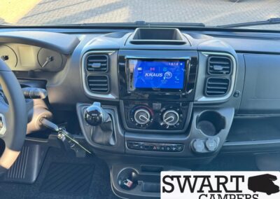 Center console and dashboard of a camper van: left-hand steering wheel, manual gear shift, round climate dials, and a glossy touchscreen displaying KNAUS. Cup holder nearby; Swart Campers watermark is visible.