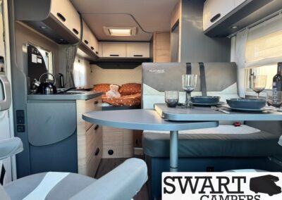Interior of a modern camper van: kitchenette left, curved dining table set for four, bed with orange bedding and a teddy in the background, SWART CAMPERS logo in corner