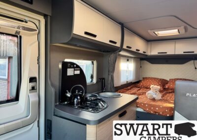 Camper van interior with kitchenette: two-burner stove and kettle, overhead cabinets, and a bed with brown polka-dot duvet and a teddy bear nearby.