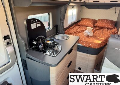 Interior of a compact camper van showing a two-burner stove, kettle, and a sink cover with a bed in orange polka-dot bedding and a teddy bear in the background.