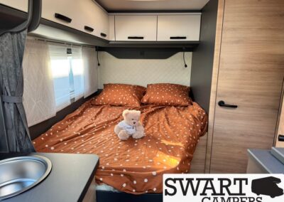 Compact camper bedroom with a double bed in rust-orange polka-dot bedding, a teddy bear, overhead storage, and a window with white curtains.