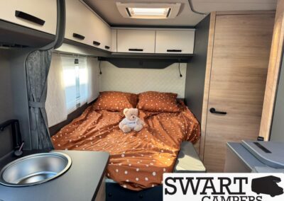 Camper van bedroom with a full bed dressed in orange polka-dot bedding and a teddy bear, above-the-bed cabinets, and a small window with curtains.
