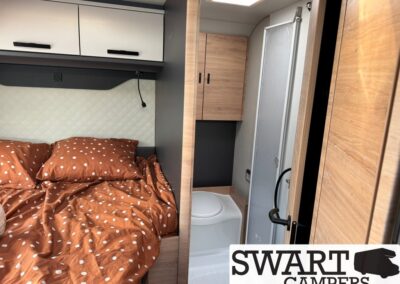 Camper van interior: bed with rust-brown polka-dot bedding, white overhead cabinets, and a compact bathroom with toilet and shower door ahead. Swart Campers interior view showing a cozy sleeping area and small washroom.