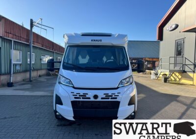 Front view of a white KNAUS camper van parked in an industrial yard with buildings on both sides and a SWART CAMPERS logo in the bottom right corner.