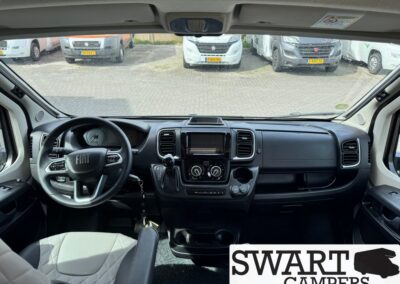 Interior view of a Fiat camper van cabin showing the steering wheel, gear selector, infotainment screen, and dashboard with SWART CAMPERS logo in the corner; outside view of parked vans through windshield.