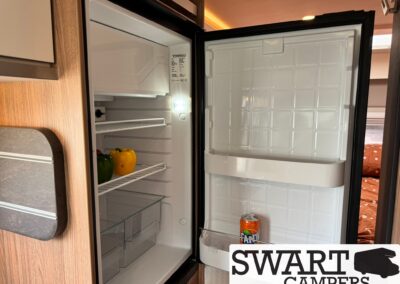 Open compact camper fridge with green and yellow bell peppers on the shelf and a can in the door rack, wood cabinetry surrounding it.