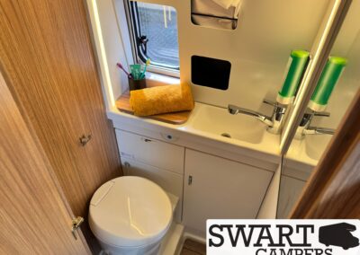 Compact camper bathroom with a white toilet, white sink, chrome faucet, a yellow towel on the counter, and a toothbrush cup by the window.