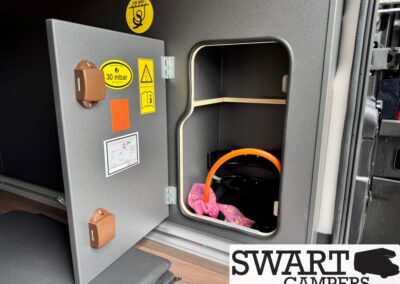 Open camper-van storage safe with warning stickers and brown latches on the door; orange securing rod and pink cloth inside.