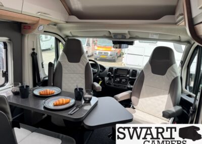 Interior of a camper van with a dining table set for two, featuring two plates with croissants, glasses, and cutlery, near the driver's and passenger's seats. The cockpit and front windshield are visible ahead.