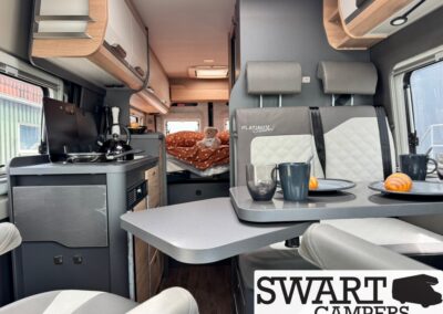 Interior of a camper van with a small kitchen on the left, a table set for two with plates and mugs, and a bed with an orange blanket and teddy in the rear. Swart Campers branding visible on seat backs.