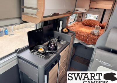 Compact camper kitchen with a two-burner stove and kettle, next to a bed with orange polka-dot bedding and a teddy bear.
