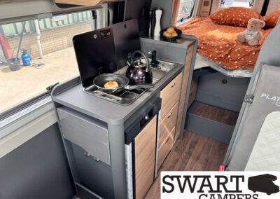 Camper van kitchen with a two‑burner stove, egg in a pan, and a kettle, plus a bed area with orange bedding and a teddy bear in the back corner. Swart Campers logo visible.