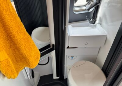 Compact camper bathroom with white toilet, small sink, and window; bright orange towel hangs nearby.