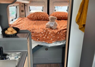 Camper interior with a bed at the rear, orange polka-dotted bedding, and a teddy bear on the bed; small kitchenette with eggs in a basket on the left.