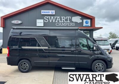 Black camper van parked outside a Swart Campers dealership with a red-roofed gray building and signage above the entrance.