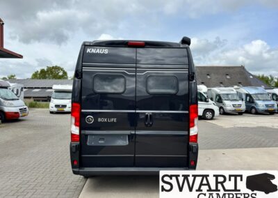 Rear view of a dark blue Knaus Box Life camper van parked in a lot, cloudy sky overhead, with other campers in the background.