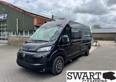Black camper van (Knaus) parked on a paved lot next to an industrial building, showing a glossy exterior and side windows.