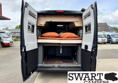 Rear of a black camper van with doors open, showing a raised bed with orange polka-dot bedding and storage space underneath.