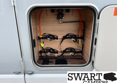 Interior of a camper storage bay with four bike-rack straps and orange propane line, seen from above in a parked trailer.