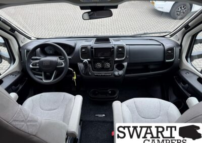 Front interior of a camper van, centered dash with touchscreen, steering wheel, and two gray front seats.