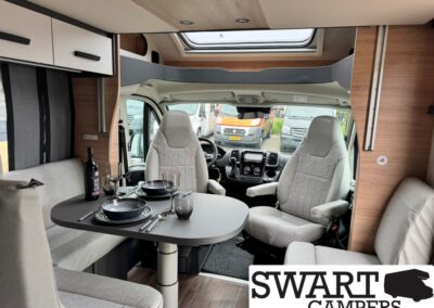 Interior of a camper van with a grey dining table set for two, wine bottle and glasses, facing the driver cab, under a skylight.