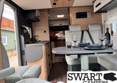 Interior of a modern camper van with a four-seat dining table set with plates and glasses, a compact kitchen, and a rear bedroom visible in the back.