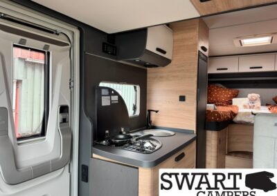 Compact camper van kitchenette with a two-burner stove, sink, and wood cabinetry, with a cozy sleeping area visible in the rear.