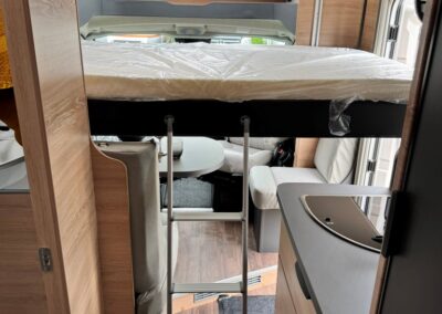 Interior of a camper van with a loft bed above a seating area, access ladder, and a kitchenette along the right wall under a skylight and window