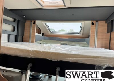 Interior of a compact camper van with a wrapped mattress on a raised loft bed over a seating/dining area and a skylight window.