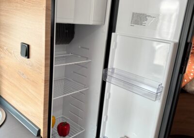 Open camper fridge with empty shelves, a yellow pepper, a red bell pepper on the lower rack, and an orange can in the door shelf.
