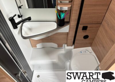 Compact camper bathroom with a white sink, black faucet, mirrored backsplash, toothbrush holder, and wooden cabinetry beside a small toilet surface.