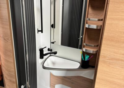 Compact camper bathroom with white sink, black faucet, and wooden storage cabinet; toothbrush cup on the counter and vertical paneling visible.