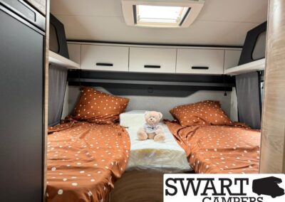 Twin bed bunk in a camper van with orange polka-dot sheets, a plush teddy bear, and overhead storage cabinets under a skylight.