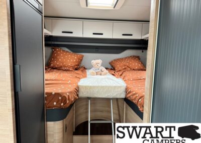 Camper bedroom with two twin bunks in orange polka-dot bedding, a teddy bear on the center mattress, and a small ladder in front.