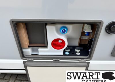 Open camper service hatch showing a white water tank with a blue fill cap and a red circular valve, plus cabinetry nearby.