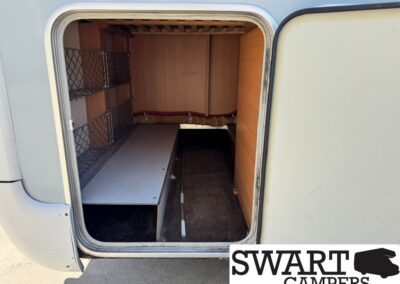 Open camper storage bay with wooden walls, metal wire shelves on the left, and a dark floor area at the back.