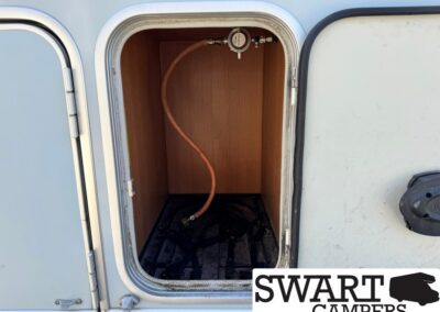 Open camper storage bay showing an orange flexible hose connected to a regulator at the top and lying inside the cabinet.