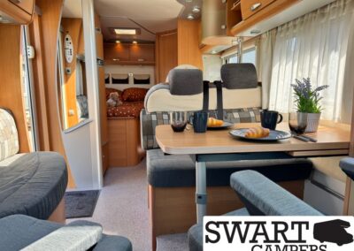Cozy camper van dining area with a table set for two: croissants on plates, dark mugs, and a potted plant by the window.