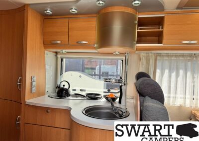Wooden camper van kitchen with curved white countertop, sink, kettle on the stove, and overhead cabinets beside a window.