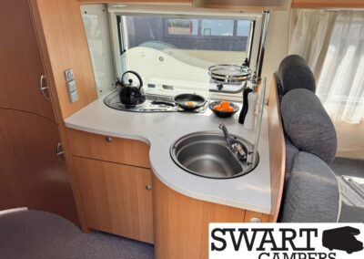 Camper kitchen with a round stainless sink, kettle on a two-burner stove, fruit bowl, and a curved white counter beside a seating area.