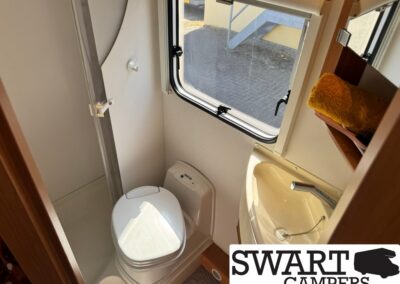 Compact RV bathroom with toilet, corner sink, and a window letting in light; a yellow towel sits on a shelf above the sink.