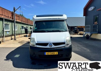 White Renault camper van with a raised KNAUS cab roof, parked in a paved lot beside industrial buildings; SWART CAMPERS logo visible in the bottom-right corner.