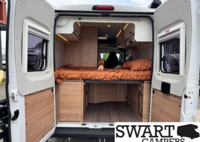 Rear of a white camper van with a raised bed, brown polka-dot bedding, and wooden storage cabinets; a small plush toy sits on the bed.
