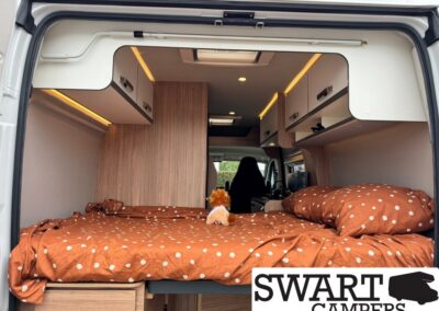Camper van bedroom with brown polka-dot bedding, a small plush toy, and overhead cabinetry.