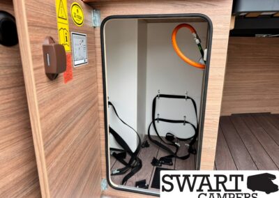 Open storage compartment of a camper with an orange hose looped at the top and black straps inside, ready for hookups.