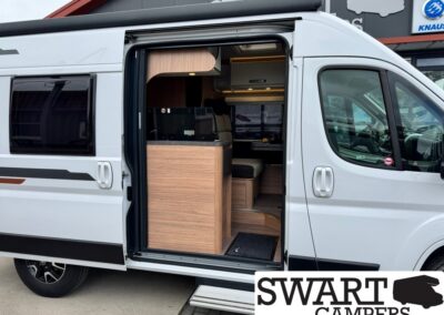 White camper van with the side door open, revealing a wooden kitchen counter and seating area inside. SWART CAMPERS sign visible outside.