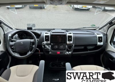 Front cabin of a campervan showing steering wheel, gear shift, center touchscreen, and dashboard controls; beige seats and cup holders visible. Parking lot seen through the windshield with other vehicles.
