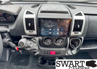 Vehicle cabin interior with a large central touchscreen, manual gear shift, climate control dials, and cup holders; SWART CAMPERS logo in the corner.