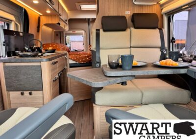 Interior of a modern camper van: a booth-style dining area with a curved table set for two, croissants and mugs on plates, plus a kitchenette with drawers and a kettle on the counter; bed and warm wood cabinetry visible in the back, soft lighting.