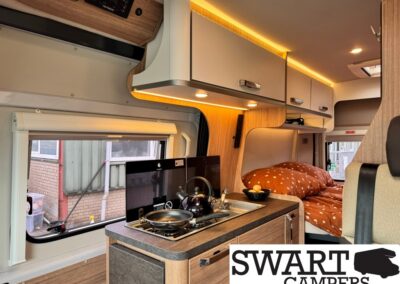 Cozy camper van interior with kitchenette (two-burner stove and kettle), overhead cabinets, and a bed with orange polka-dot bedding in the back.